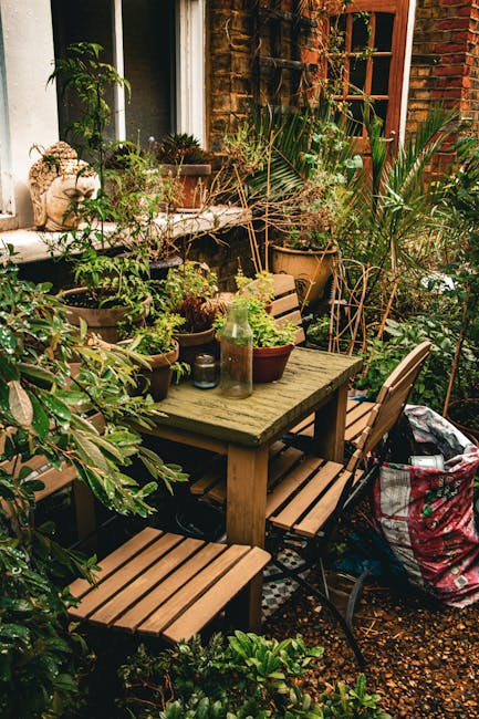 A cozy urban garden patio featuring a weathered wooden table surrounded by matching wooden and metal chairs, set against a brick wall with a window. The table is decorated with small potted plants, in