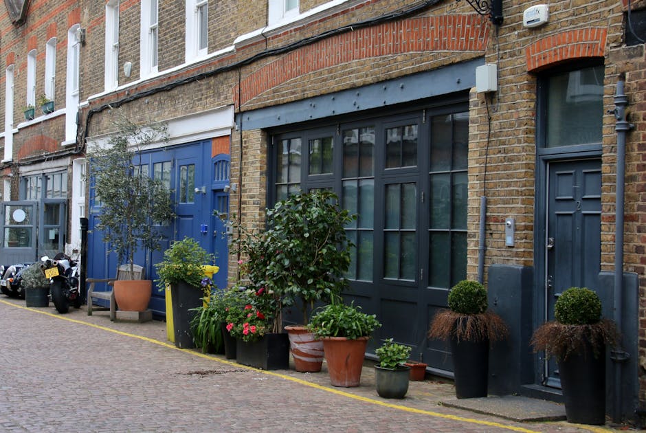 A narrow Stoke Newington mews features a row of terraced brick houses with variously colored doors and window frames, including dark gray and blue. In front of the houses, there is a small urban garde